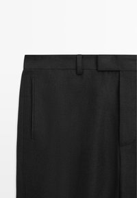 Pantalon noir coupé sur mesure avec passants de ceinture, poche latérale fendue et une ceinture plate, visible de la taille au haut de la cuisse.