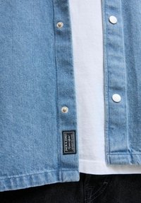 Lichtblauwe denimshirt met zilveren drukknoopjes, gelaagd over een wit shirt. Bevat een zwart merketiket op de zoom, met een gladde textuur.