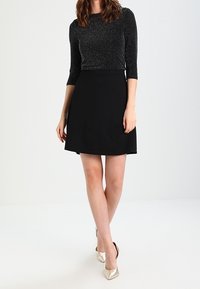 Černá krátká sukně v kombinaci s třpytivým černým topem s dlouhým rukávem. Modelka má na sobě metalické boty s špičatou špičkou. Jednoduchý design, hladká textura.