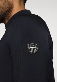 Tengerészkék, hosszú ujjú ing, sima textúrával. Bal vállán fekete EAZ logóval ellátott folttal. Klasszikus galléros dizájn.