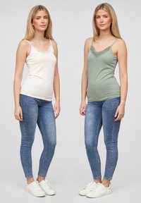 To kvinner iført tettsittende tank topper; den ene i lys krem med blonderkant, den andre i oliven grønn. Begge kombinert med blå smale jeans og hvite sko.