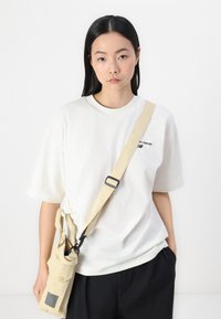 T-shirt en coton blanc avec un col rond, associé à un sac bandoulière beige avec un clip, une poche et une sangle plate. Pantalons noirs.