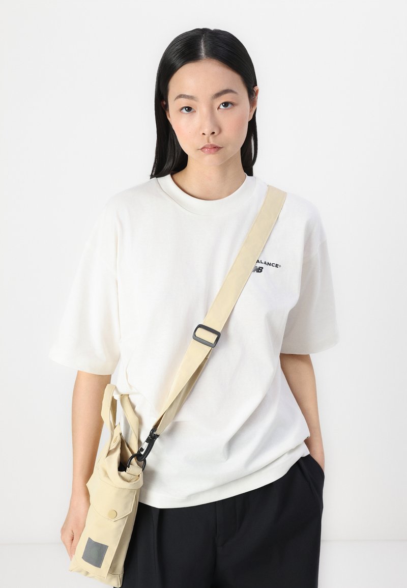 Weißes Baumwoll-T-Shirt mit Rundhalsausschnitt, kombiniert mit einer beigen Crossbody-Tasche mit Clip, Tasche und flachem Trageriemen. Schwarze Hose.