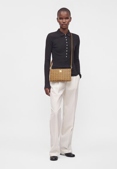 Femme portant un haut noir à manches longues, un pantalon blanc à jambes larges, des chaussures noires, portant un sac bandoulière marron avec un motif.