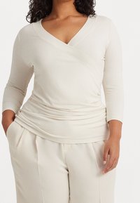 Ljusbeige omslagstopp med djup V-ringning och trekvartslånga ärmar, som har en draperad textur och ett mjukt tyg. Matchas med ljusa byxor.