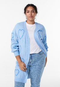Svetlo modrý kardigán s veľkými vzormi srdca, vyrobený z mäkkého materiálu. Má otvorený predný diel a rebrované manžety. Nosený cez bielu košeľu a modré džínsy.