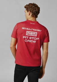 Červené bavlněné tričko s krátkými rukávy. Na zadní straně je bíly text: "RED BULL RACING", "RBR" a "PIT STOP CREW". Jednoduchý, neformální design.