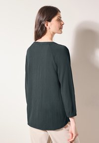 Blusa textured di un verde scuro, con maniche lunghe e orlo arrotondato. Presenta un semplice collo a giro e dettagli di cucitura discreti.