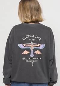 Sudadera gris con un colorido estampado gráfico de un pájaro y montañas, con el texto "Vida Eterna", "Sociedad Kaotiko" y un símbolo ankh.