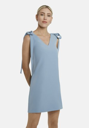 Frau trägt ein ärmelloses hellblaues Kleid mit gebundenen Schleifen an den Schultern, steht vor einem schlichten weißen Hintergrund.