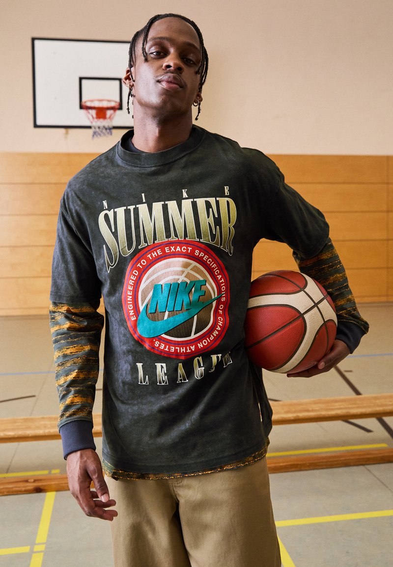 Nike grafisk langermet skjorte med en falmet svart farge, med teksten "SUMMER" og et turkis logo; holder en basketball, iført khaki-bukser.