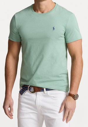 Man draagt een lichtgroen t-shirt met korte mouwen met een klein blauw logo, witte broek, gestreepte riem en een bruin polshorloge.