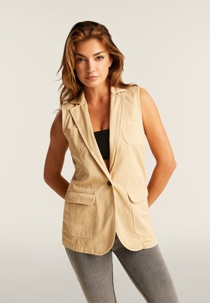 Mujer con cabello castaño claro, vestida con blazer beige sin mangas, top corto negro y jeans grises, de pie frente a un fondo claro y liso.