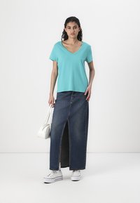 Turkise v-hals t-shirt parret med en lang denimnederdel med en slid foran. Modellen bærer også en hvid håndtaske og har hvide sneakers på.