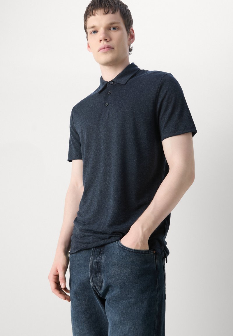 Dunkelgraues Poloshirt mit Kragen, drei Knöpfen und kurzen Ärmeln; aus einem weichen, strukturierten Stoff gefertigt; kombiniert mit blauen Jeans.