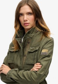 Grøn militærjakke i stil med lynlås Krave, brystlommer og et patch med "SUPERDRY." Struktureret stof med et robust design.
