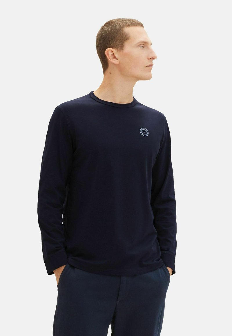 Marineblaue Langarm-T-Shirt mit runder Halslinie, versehen mit einem kleinen gestickten Logo auf der Brust und einer glatten, weichen Textur.