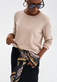 Beżowy sweter dziany z okrągłym dekoltem, w zestawieniu z wzorzystym czarno-złotym szalikiem, charakteryzującym się misternymi wzorami i miękką teksturą.