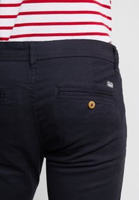 Granatowe spodnie o gładkiej teksturze, z dwoma tylnymi kieszeniami i drewnianym guzikiem jako detalem. Na pasku widoczna jest mała metka marki.