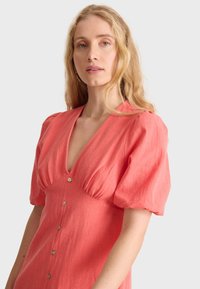 Koralfarbenes Hemdkleid mit Puffärmeln, tiefem V-Ausschnitt und strukturierter Stoffqualität. Es hat ein tailliertes Oberteil und eine A-Linie Silhouette.