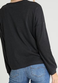 Nainen, jolla on päällään löysä tummanharmaa pitkähihaine paita ja vaaleansiniset farkut, näkyy takaa neutraalia taustaa vasten.