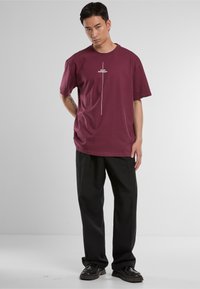 Maroon oversized t-shirt made of cotton. Features a white vertical graphic saying "Think Different." Paired with black trousers and shoes.