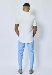 Chemise blanche à manches courtes avec un ourlet arrondi, associée à un pantalon slim-fit bleu clair et des chaussures grises, vue de dos.