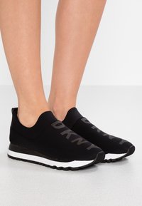 Baskets noires à enfiler en tissu extensible avec une texture lisse, dotées d'une semelle rayée noire et blanche et d'un grand logo.