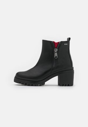 Bottine noire à talon épais, fermeture éclair latérale partiellement ouverte révélant une doublure intérieure rouge, et semelle crantée.