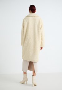 Krämfärgad fake pälsjacka med en plyschliknande textur, oversize passform och bred krage. Bärs över en beige klänning med en mittlång skåra.
