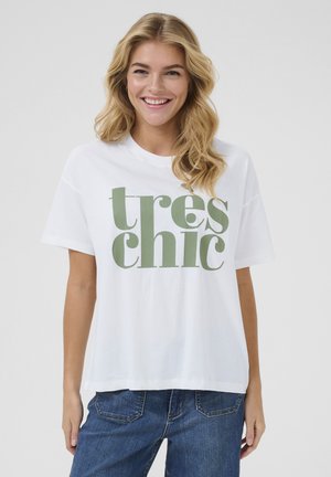 Vit bomullströja med översized text i grönt som säger "tres chic." Korta ärmar med en avslappnad passform, i kombination med blå denimjeans.