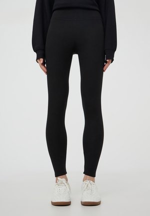 Persona che indossa leggings neri aderenti e sneakers bianche, in piedi contro uno sfondo chiaro e semplice con le mani rilassate ai lati.