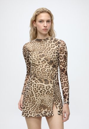 Jeune femme aux cheveux blonds portant une mini-robe à manches longues imprimée léopard avec une fente latérale, debout devant un fond blanc uni.