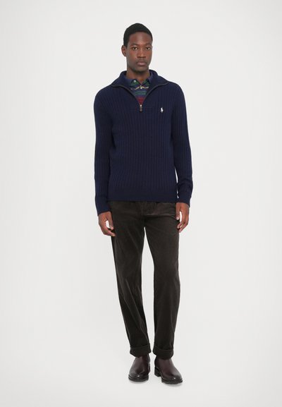 Pulover tricotat cu model in dungi navy, cu guler cu fermoar la un sfert, asortat cu pantaloni din corduroy maro și cizme negre până la gleznă. Logo discret pe piept.