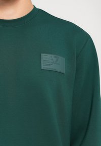 Sötétzöld pulóver, bal mellen hímzett foltlogóval, amelyen az "EA7 Emporio Armani" felirat olvasható.