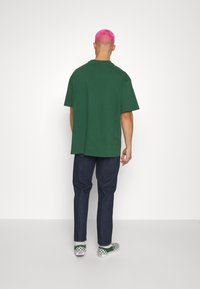Grön oversized t-shirt, korta ärmar, bomullsmaterial, bärs med mörkblå jeans, schackrutig slip-on skor och rosa buzz cut frisyr.