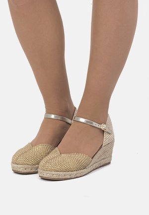 Zapatos de cuña con parte superior de fibra natural dorada tejida, puntera redonda y correa metálica en el tobillo. Suela intermedia de yute texturizado y suela de goma.