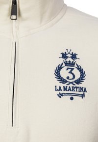 Sudadera cremosa con cremallera y un emblema bordado en azul marino que muestra dos jugadores de polo, una corona, una corona de laurel, el número 3 y el texto "LA MARTINA".