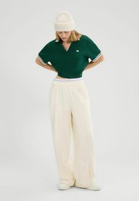 Groene cropped polo met witte accenten, gecombineerd met crèmekleurige wijde broek en een lichtbeige gebreide beanie. Zachte stofstructuur.