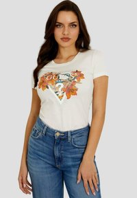 T-shirt en coton blanc avec un graphique triangulaire orné d'accents floraux et de détails métalliques, associé à un jean bleu taille haute.