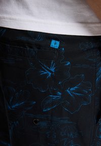 Czarne szorty z tkaniny z niebieskim motywem kwiatowym, pojedyncza kieszeń z tyłu oraz mała niebieska metka marki na pasku.