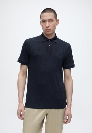 Ung mann iført en strukturert svart kortermet poloskjorte og beige bukser, står foran en ensfarget lys bakgrunn.