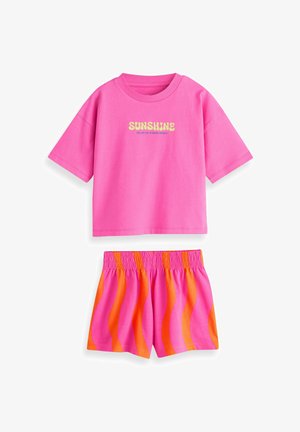 Roza bombažna majica s potiskom "SUNSHINE" v rumeni barvi, združena z kratkimi hlačami z elastičnim pasom, okrašenimi z rožnatimi in oranžnimi valovitimi črtami.