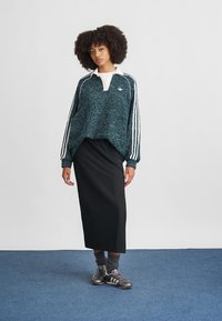 Sweater oversized texturizado verde com listras brancas, colarinho branco e um logótipo bordado, combinado com uma saia midi preta e ténis escuros.