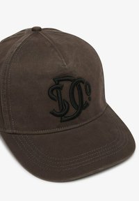 Gorra de béisbol marrón hecha de tela, con un logo negro bordado en la parte frontal. Forma redondeada con un visera curvada y costuras visibles.
