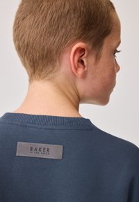 Marineblauer Sweatshirt mit einem gerippten Halsausschnitt, das auf der Rückseite ein graues Lederetikett mit der Aufschrift "BAKER by TED BAKER" trägt. Weiche Textur, legeres Design.