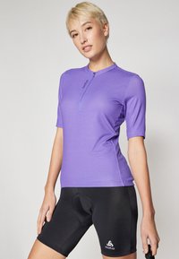 Lilla kortermet sykkeltrøye med glidelås i kragen, laget av lett materiale, kombinert med svarte polstrede sykkelshorts med logo.