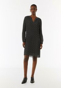 Schwarzes gemustertes Kleid mit langen Ärmeln, plissiertem Rock und V-Ausschnitt, aus leichtem Stoff. Kombiniert mit schwarzen High Heels.