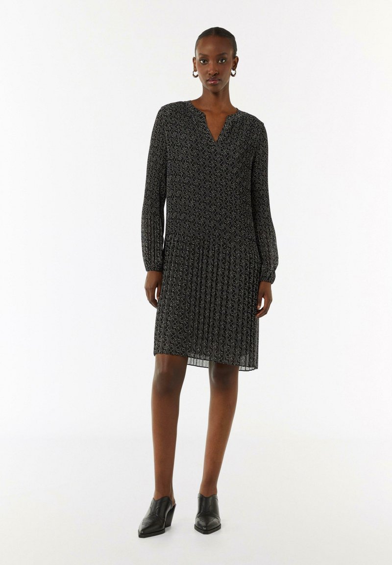 Schwarzes gemustertes Kleid mit langen Ärmeln, plissiertem Rock und V-Ausschnitt, aus leichtem Stoff. Kombiniert mit schwarzen High Heels.