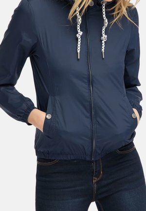 Frau trägt eine marineblaue Jacke mit Reißverschluss und weißen Kordeln, mit Knopfverschlusstaschen, elastischen Bündchen und dunkelblaue Jeans, Hände in den Taschen.